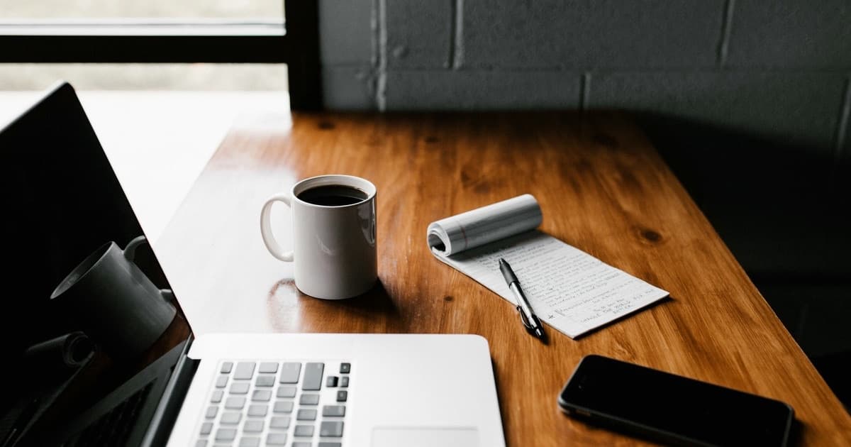 A laptop, a coffee mug, a notepad and a phone on a wooden desk