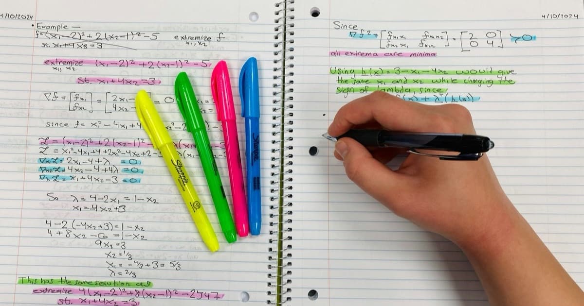 A hand writing maths notes in a spiral notebook beside four highlighters