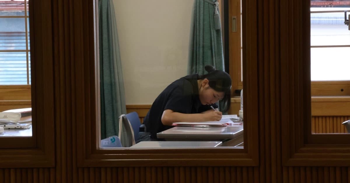 A student bent over a desk writing, seen through a classroom window